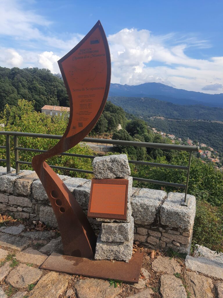 Panneau en métal orangé, de forme géométrique et oblongue, contre un muret en brique, avec en fond le paysage de montagne de la corse et le ciel bleu