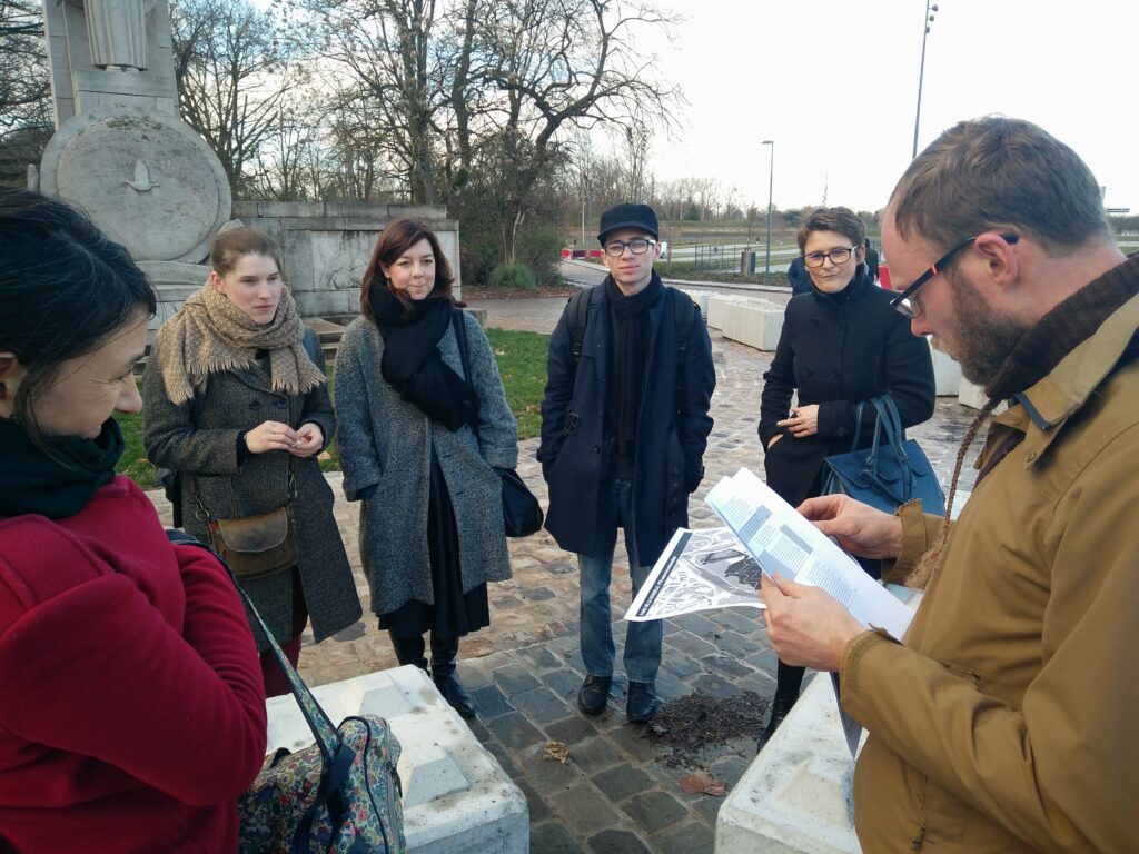 Cinq personnes écoutant une sixième personne, document en main, lors d'une visite sur un site patrimonial en plein air