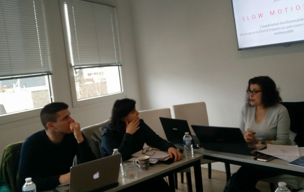 Deux personnes attablées à un bureau d'école regardent fixement une troisième personne assise à un autre bureau en train de réaliser une présentation grâce à un écran positionné derrière elle.